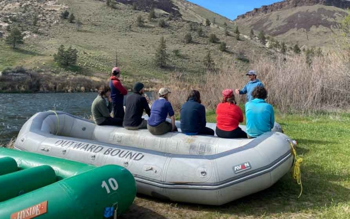 a group of gap year students rest on the side of a beached raft while listening to an instructor on an outward bound outdoor educator course